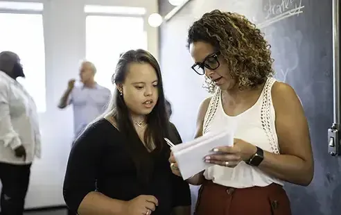 Two women standing in front of a blackboard, engaged in discussion or teaching, with educational materials visible.